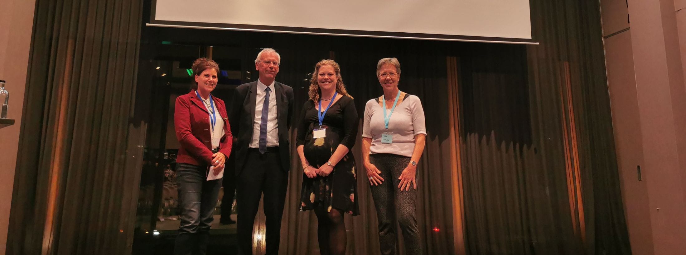 Vier personen staan op een podium voor een presentatiescherm tijdens een symposium over geriatrische revalidatiezorg.