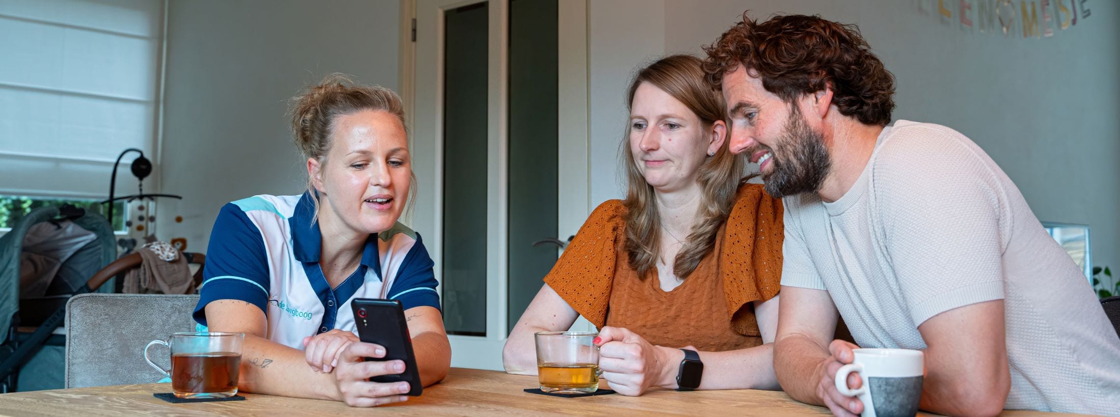 Nieuwe ouders zitten thuis aan tafel met een Zorgboog medewerker die iets op een telefoon laat zien.