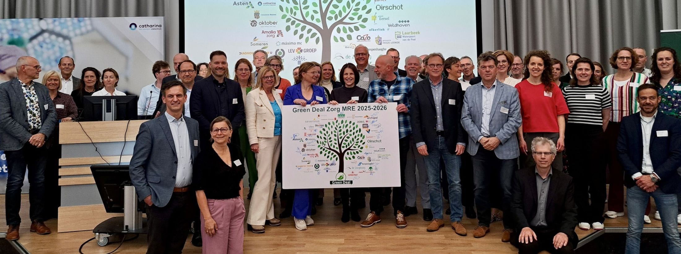 Grote groep vertegenwoordigers van zorgorganisaties poseert met een poster op een podium bij de ondertekening van de regionale Green Deal Zorg 2025–2026.