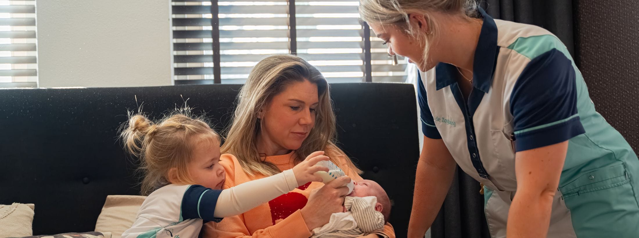 Zorgboog medewerker begeleidt een moeder en haar dochter bij het geven van een flesje aan een pasgeboren baby.