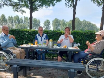 Cliënten in rolstoelen en medewerkers eten en praten samen aan een picknicktafel.