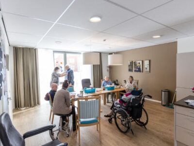 Cliënten en medewerkers zitten en staan samen aan een eettafel met kunst aan de muren.