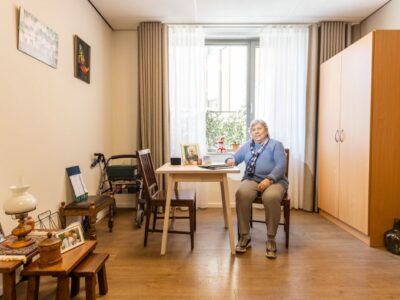 Cliënt zit aan tafel in een zonnige kamer met kasten en foto’s aan de muur.