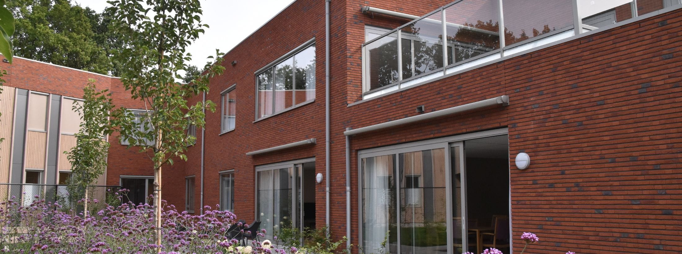 Buitenaanzicht van locatie Hoge Vogelsanck met een paarse bloemenperk op de voorgrond.