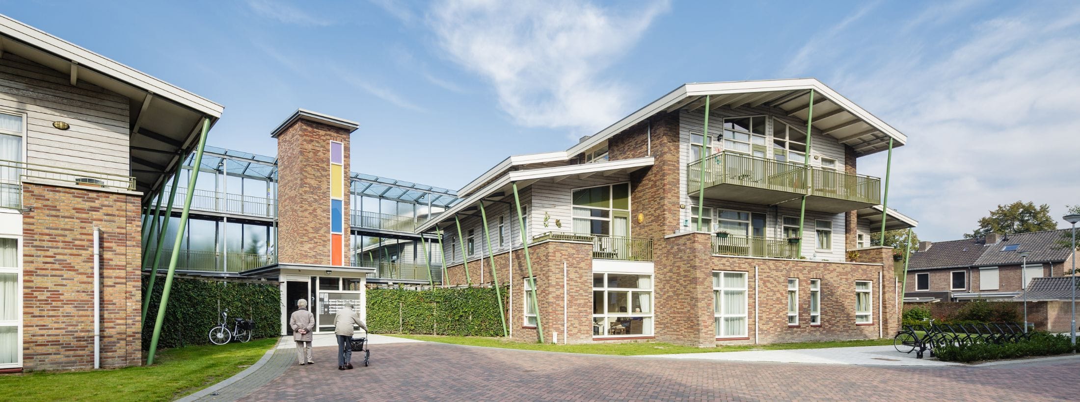 Buitenaanzicht van locatie Wellenshof met een ouder stel lopend bij de ingang.