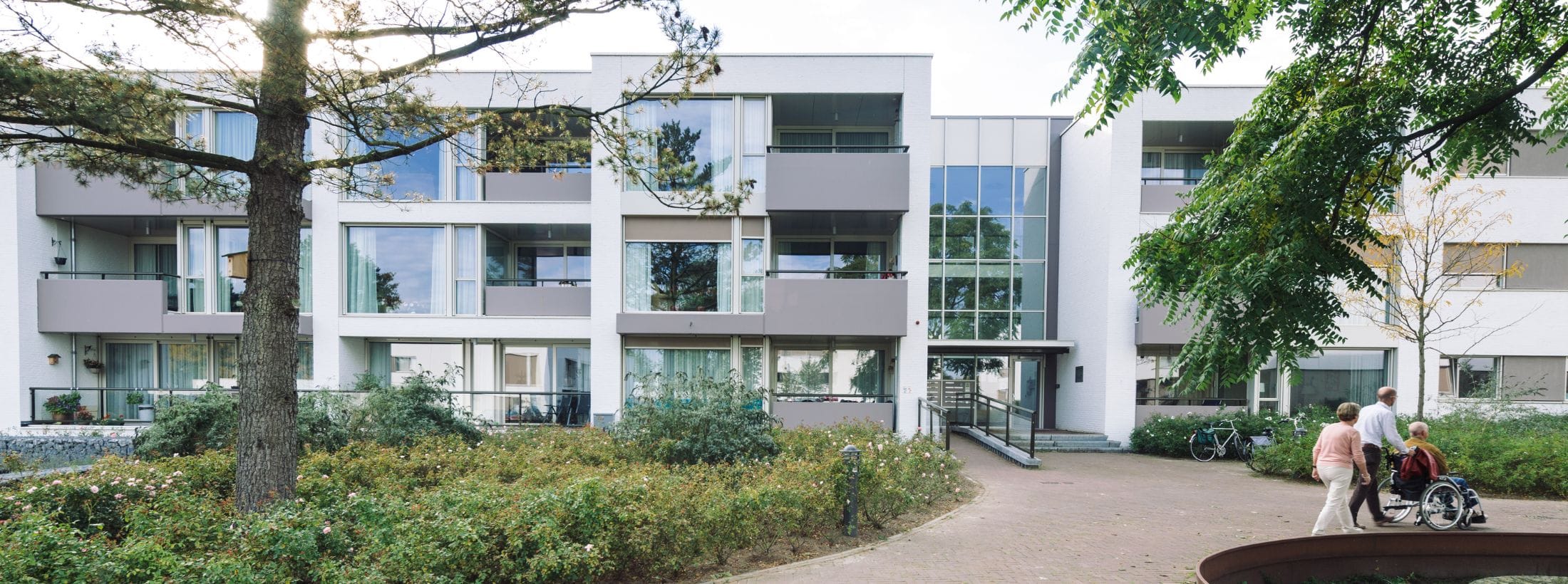 Buitenaanzicht van locatie Zonnetij met bomen en cliënten op de voorgrond.