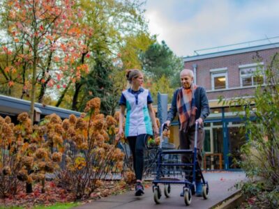 Zorgmedewerker en cliënt met rollator wandelen samen buiten.