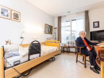Cliënt zit aan tafel in een kamer met een bed, tv en foto's aan de muur.