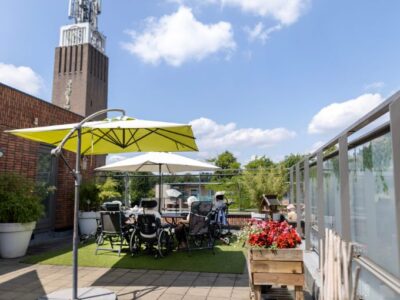 Een groep cliënten zit op het terras van locatie Someren-Eind onder parasols, met een kerk op de achtergrond.