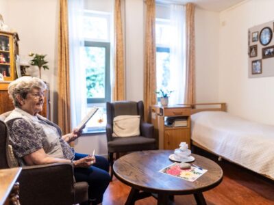 Cliënt zit in een stoel en maakt een puzzel in een kamer met een bed, tafel en gele gordijnen.