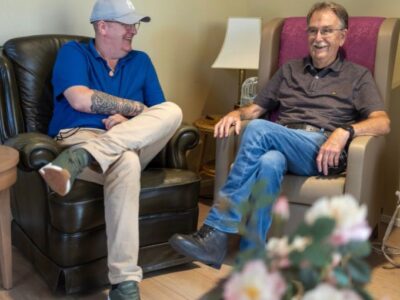 Cliënt en Zorgboog medewerker zitten naast elkaar op comfortabele stoelen en hebben een lachend gesprek.