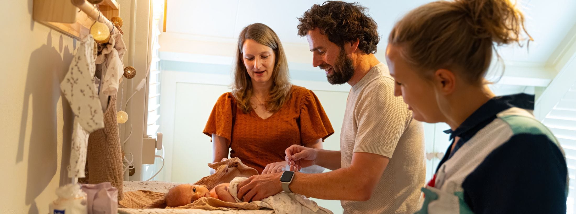 Zorgboog medewerker staat naast ouders die hun pasgeboren baby op tafel verschonen en in gesprek zijn met de medewerker.