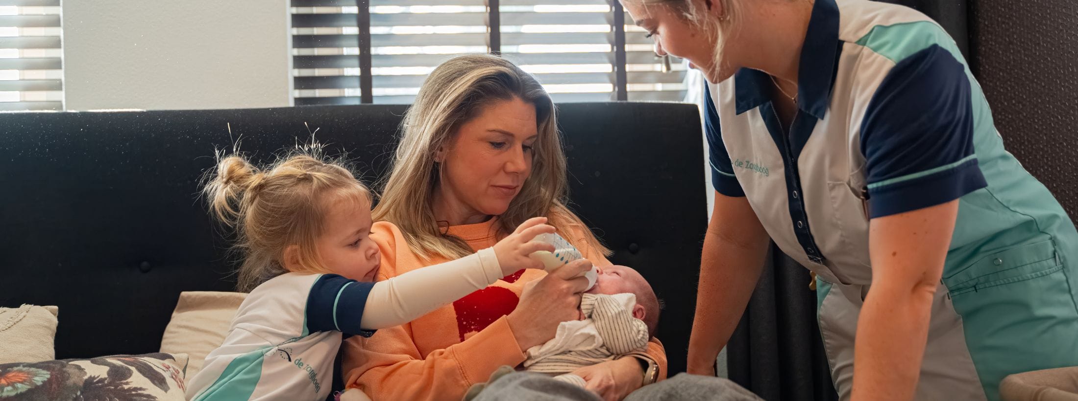 Zorgboog medewerker begeleidt een moeder en haar dochter bij het geven van een flesje aan een pasgeboren baby.